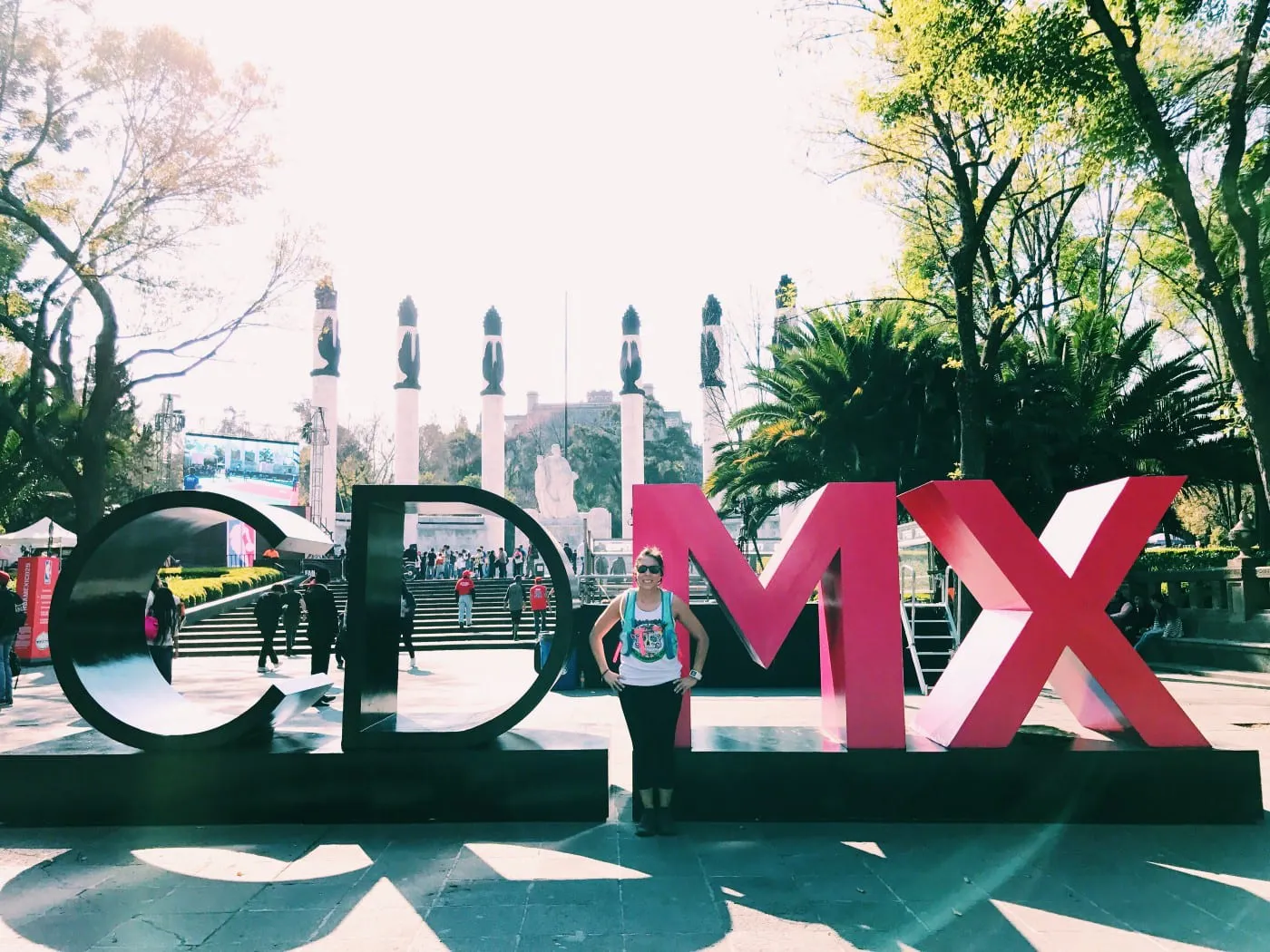 A woman in a white tank top, teal backpack and sunglasses posing in front of a sculpture in Mexico City, Mexico.