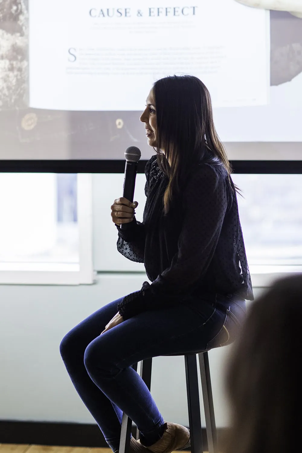 A woman sitting on a stool speaking into a microphone during a presentation.
