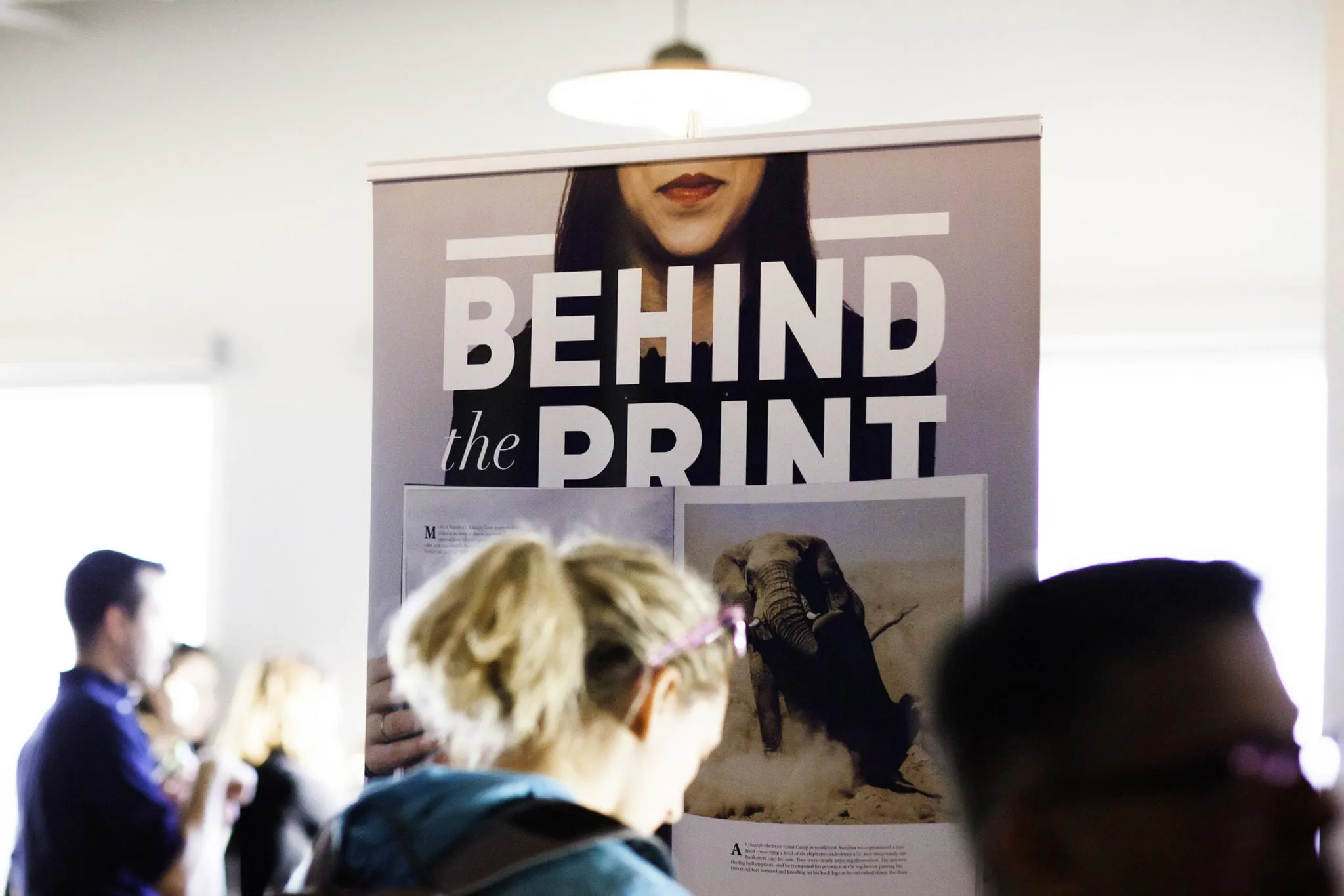 A woman standing in front of a custom hanging banner printed with Behind the Print in white.
