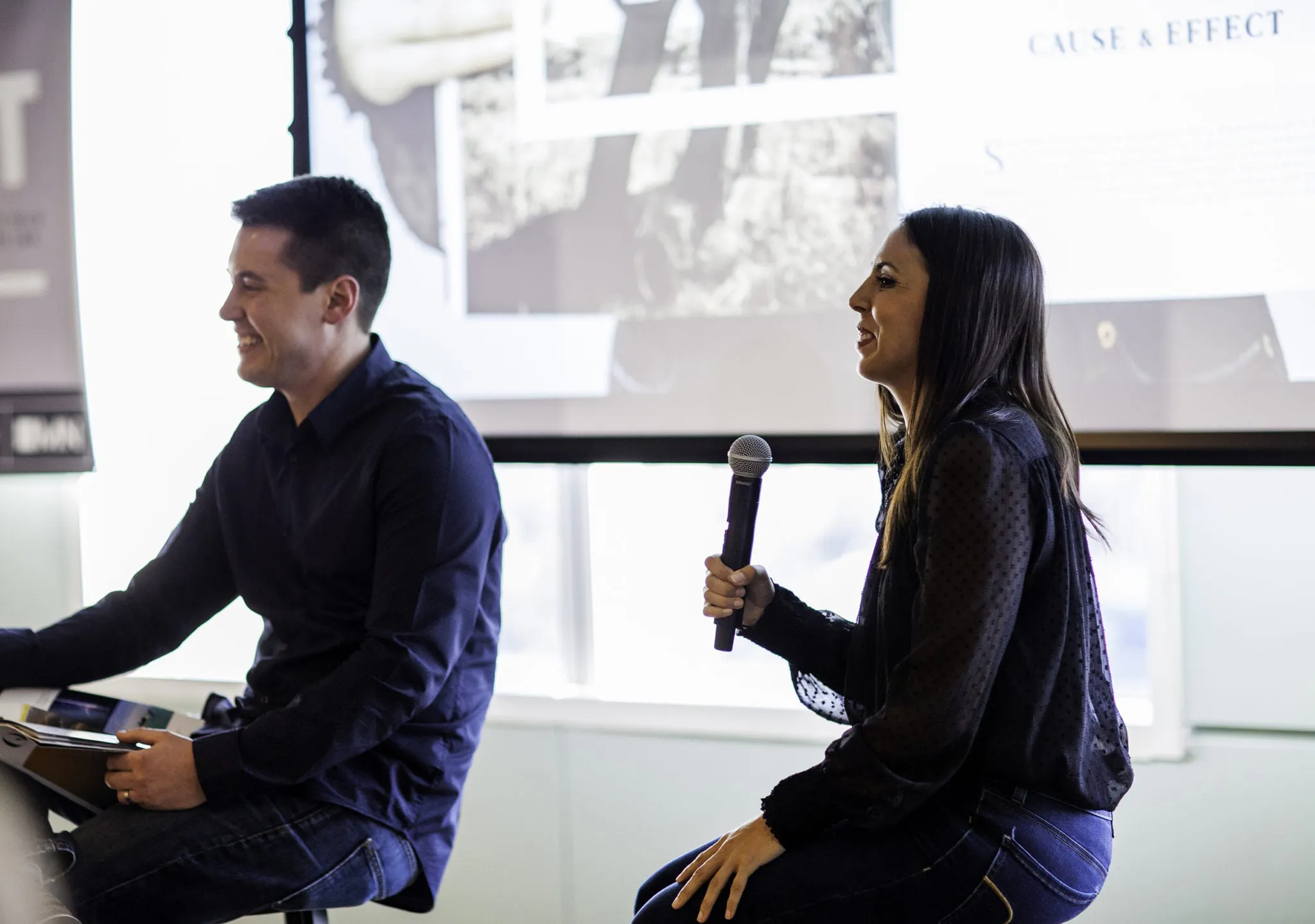 A man and a woman sitting on stools and holding microphones with a banner behind them during a presentation.