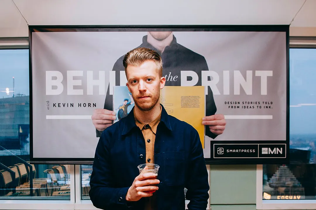 A man holding a drink in a plastic cup standing in front of a banner printed with Behind the Print.
