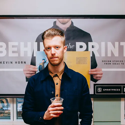 A man holding a drink in a plastic cup standing in front of a banner printed with Behind the Print.