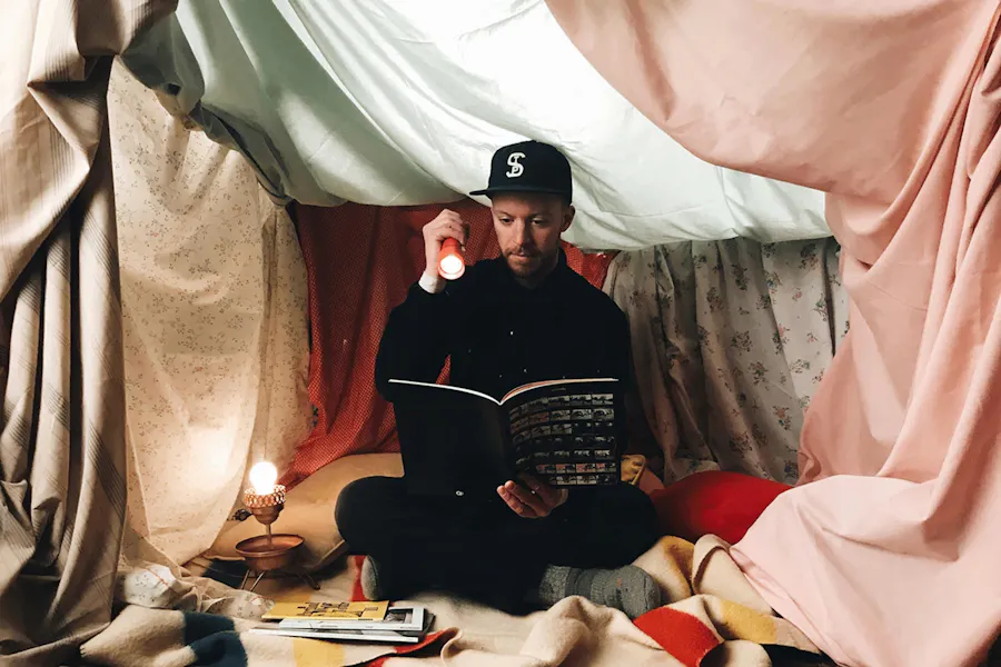 A man sitting in a blanket fort holding a flashlight and a custom booklet.