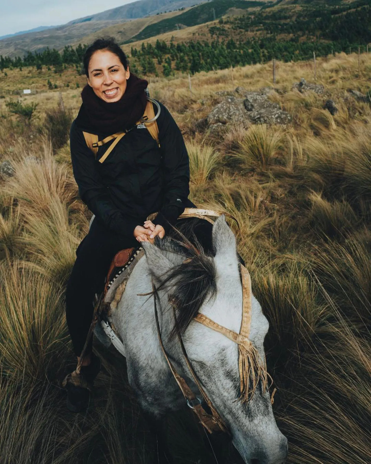 A woman wearing a black scarf and jacket smiling and riding a horse through a grassy pasture.