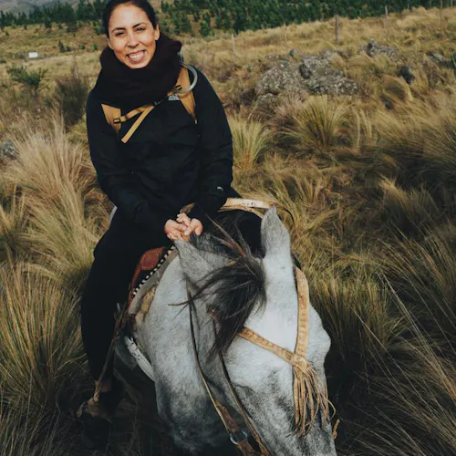 A woman wearing a black scarf and jacket smiling and riding a horse through a grassy pasture.