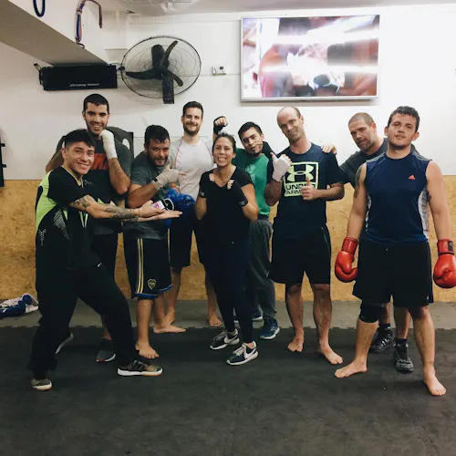 A woman smiling and standing with a group of men in a boxing gym with a fan behind them.