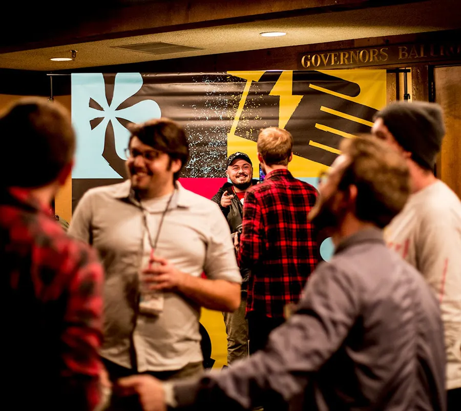 A group of men standing and talking to each other in front a step and repeat banner printed in black, yellow and blue.