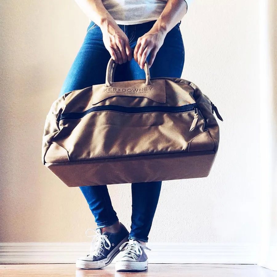 A woman in blue jeans, tennis shoes and a white top holding a brown Ker and Downey duffle bag.