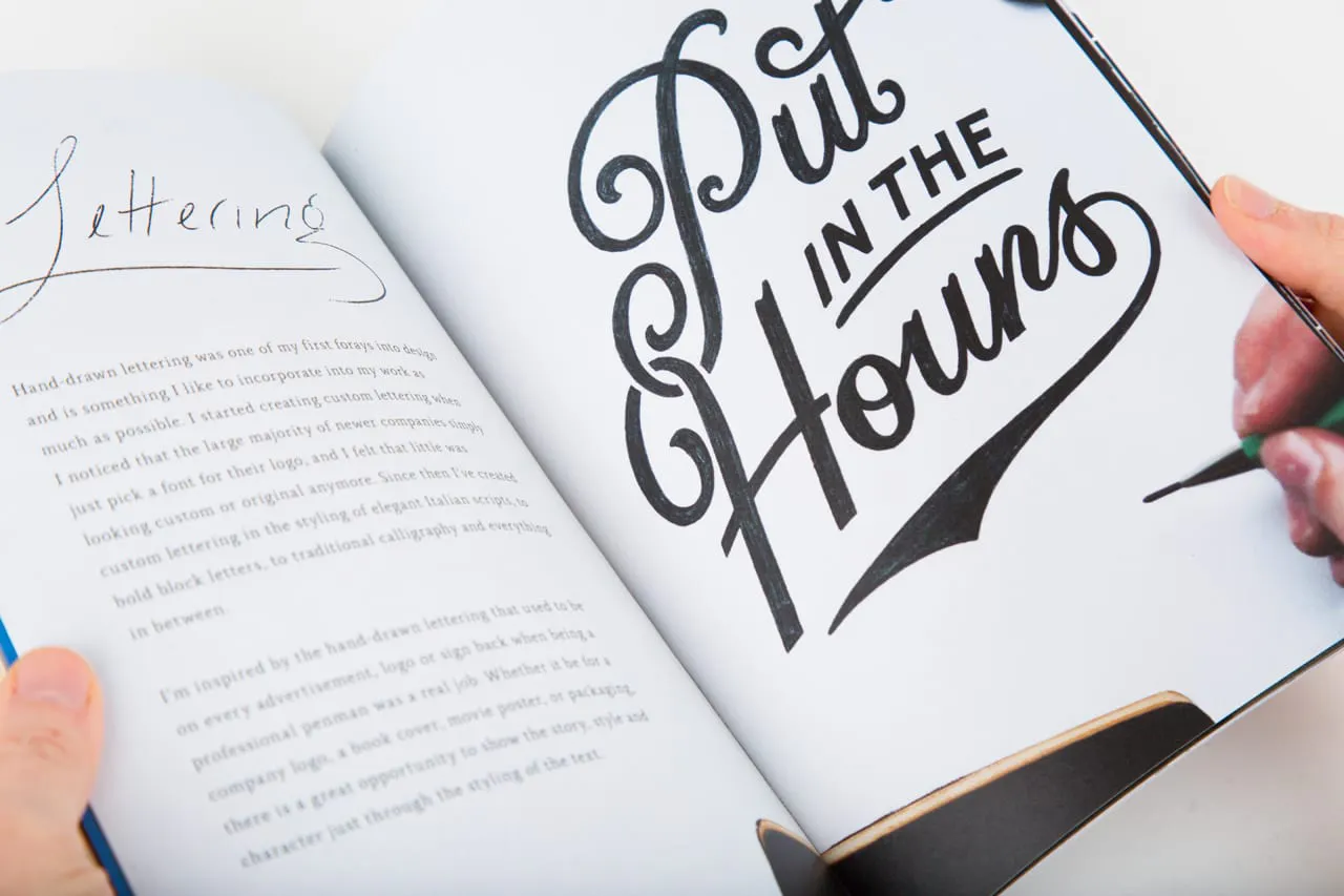 A portfolio book laying open to a print with Put In The Hours in cursive.