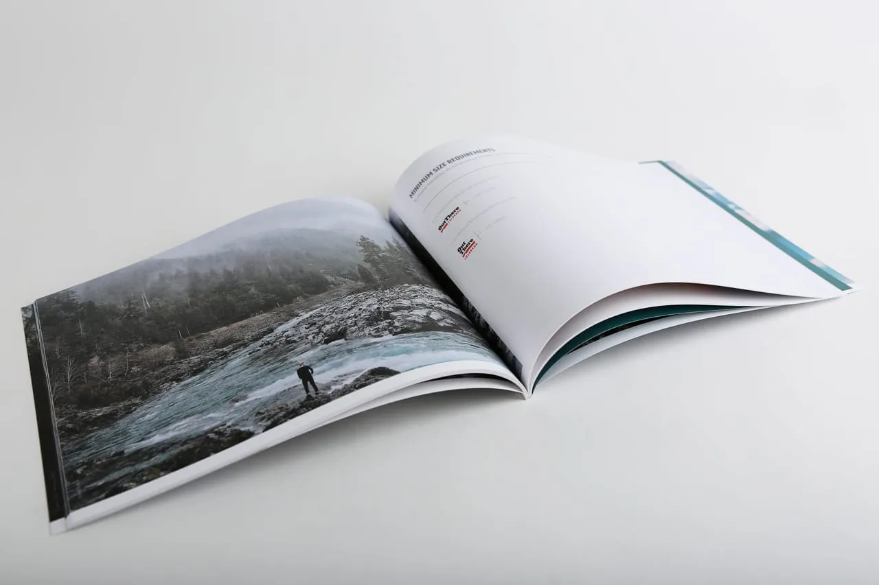 A brand manual laying open to an image of a person standing on the rocky shoreline of a winding river in a forest.