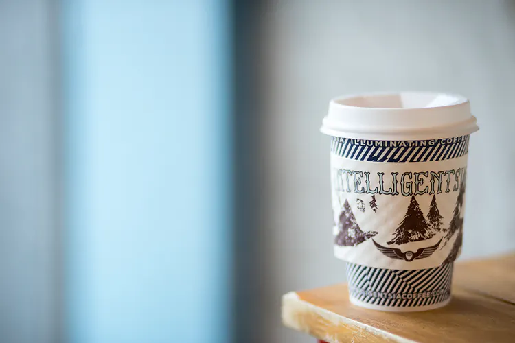 A to-go coffee cup with a sleeve printed with Intelligentsia sitting on the corner of a wooden table.