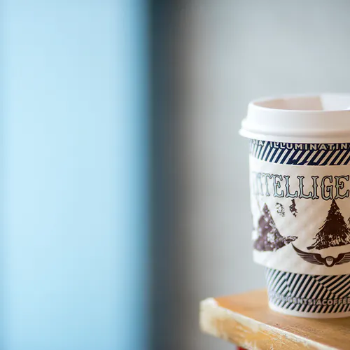 A to-go coffee cup with a sleeve printed with Intelligentsia sitting on the corner of a wooden table.