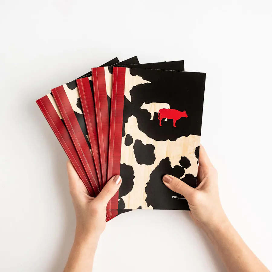 Two hands holding five fanned-out custom menus with a red spine and a cream and red cow design on the front.