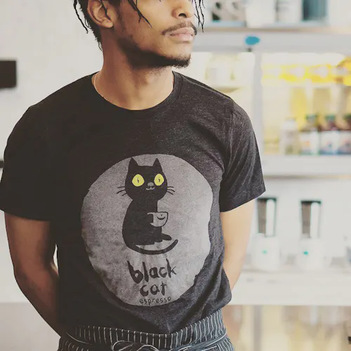 A man standing with his hands behind his back wearing a black T-shirt printed with a black cat graphic.