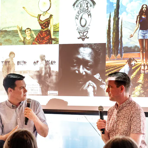 Two men sitting and holding microphones during a presentation with photography hanging behind them.