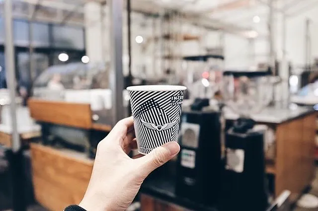 A hand holding a small coffee cup printed with a navy blue striped design in front of a coffee shop.