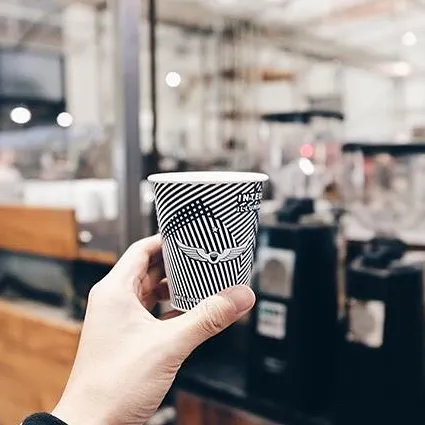 A hand holding a small coffee cup printed with a navy blue striped design in front of a coffee shop.