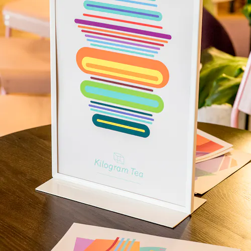 A custom art print in bright orange, blue, pink, purple and yellow in a white free-standing frame sitting on a table.