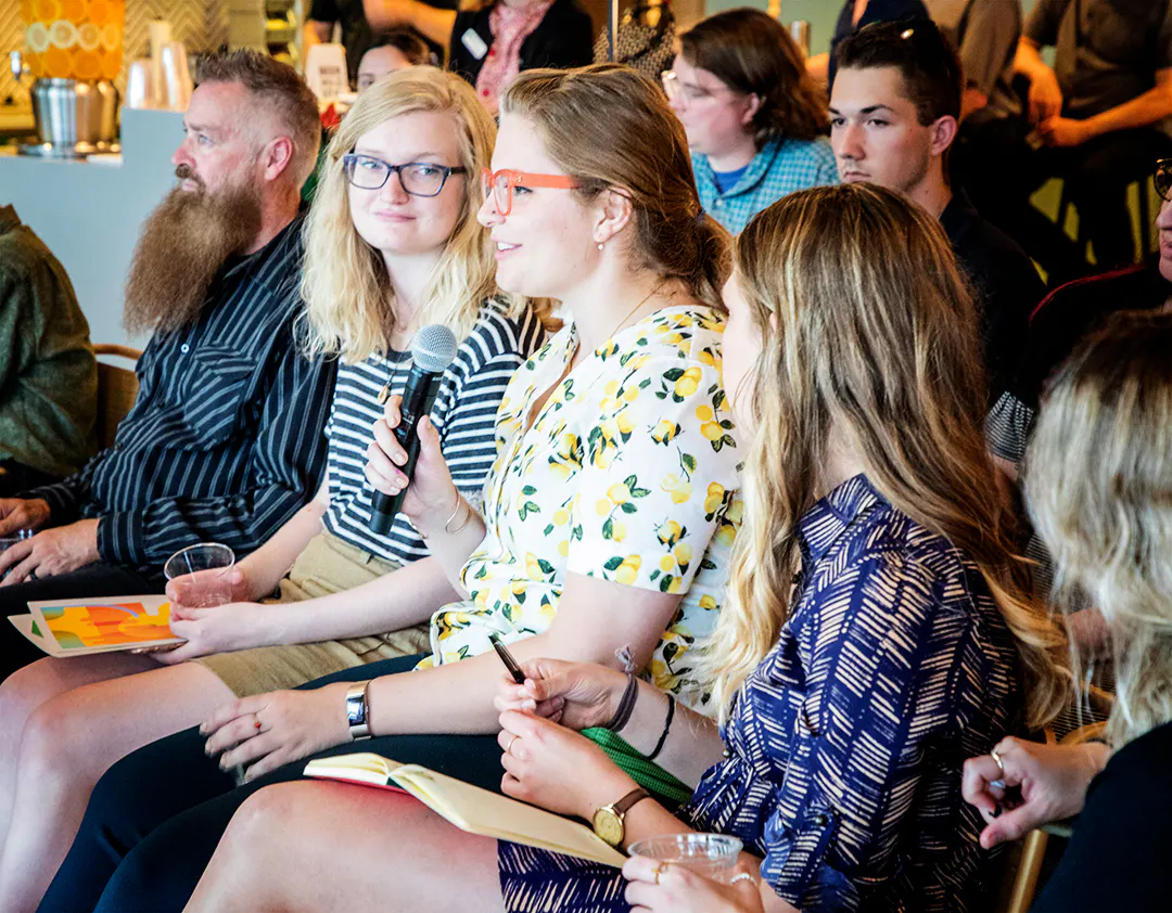 A woman with glasses holding a microphone sitting in an audience with other people sitting around her.