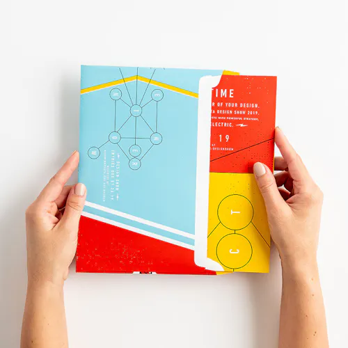 Two hands pulling a folded direct mail poster out of its envelope with a blue, red and yellow design.