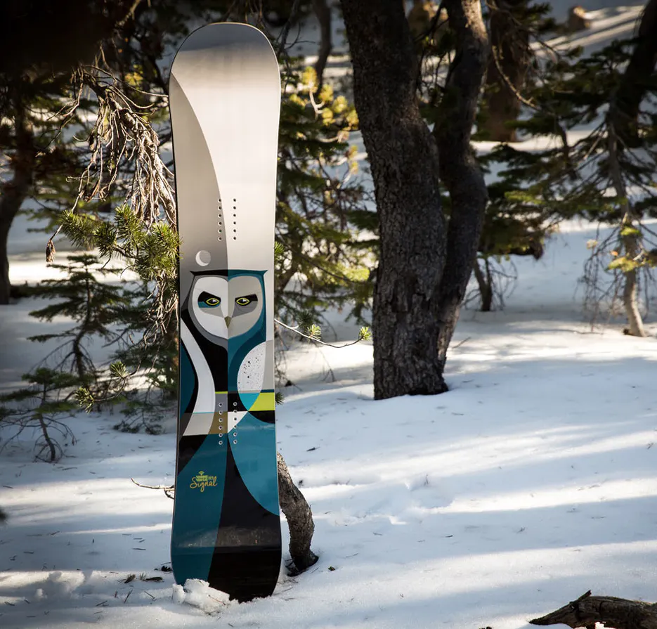 A snowboard standing in the snow with custom artwork of an owl on the bottom.