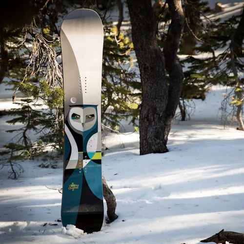 A snowboard standing in the snow with custom artwork of an owl on the bottom.