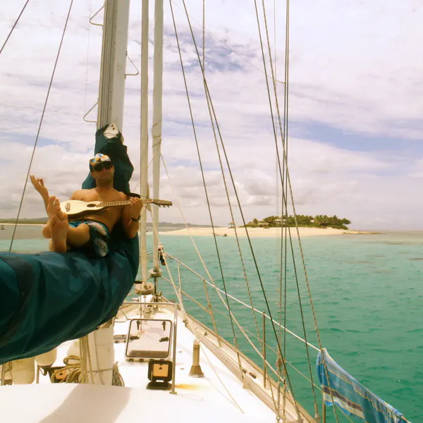 A man wearing a hat and sunglasses sitting on a rolled up sail on a sailboat in blue-green water.