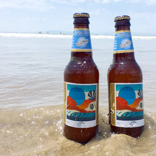 Two Blue Moon beer bottles sitting in the surf on the beach with custom labels.