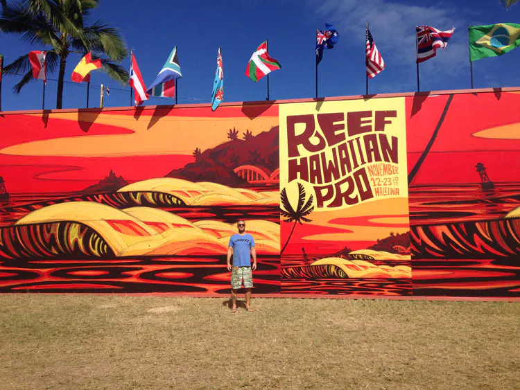 A man standing in front of a custom banner printed with a surfing design in shades of orange and red.