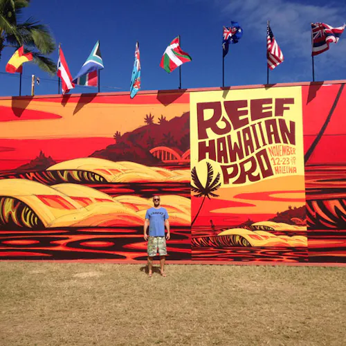A man standing in front of a custom banner printed with a surfing design in shades of orange and red.