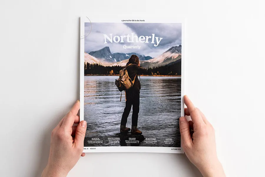 Two hands holding a perfect bound booklet with Northerly and a person standing on a rock in a lake.