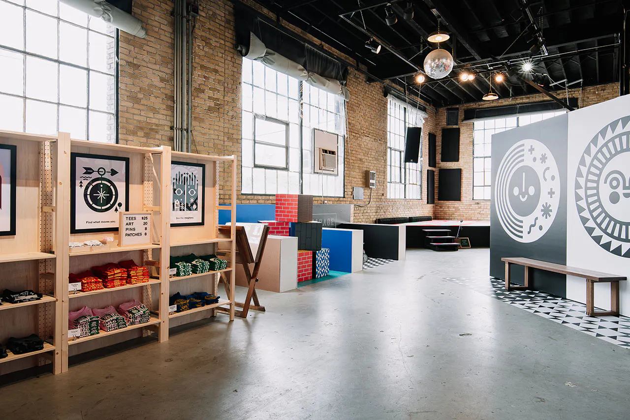 A warehouse space with brick walls, a stage in the corner, blocks with custom decals on the sides and a T-shirt display.