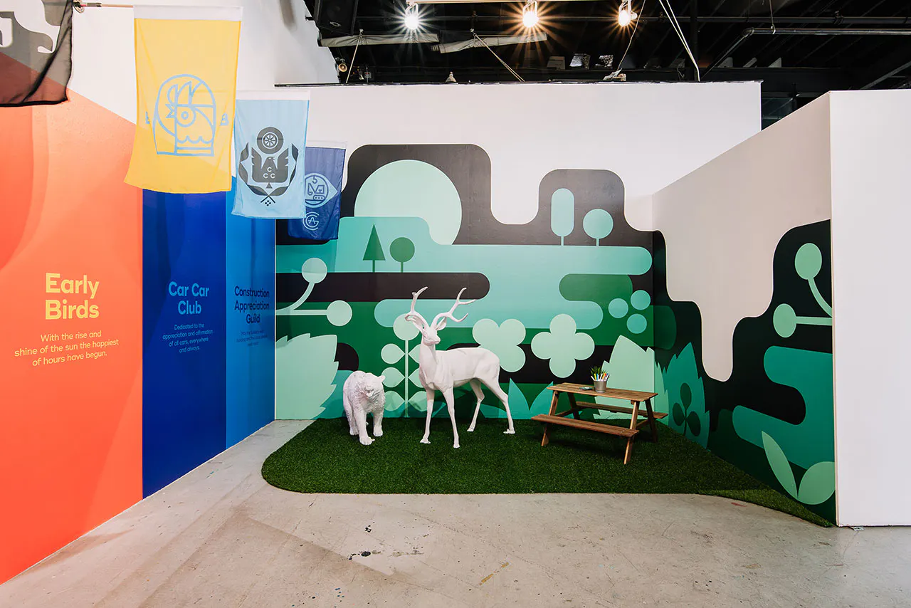 A visual display of two white animal sculptures and a small picnic table on fake grass with a green wall decal behind it.
