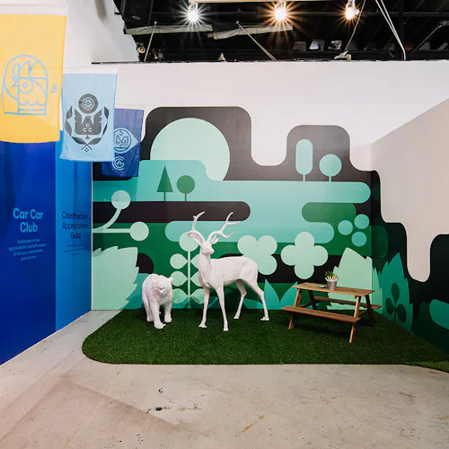 A visual display of two white animal sculptures and a small picnic table on fake grass with a green wall decal behind it.