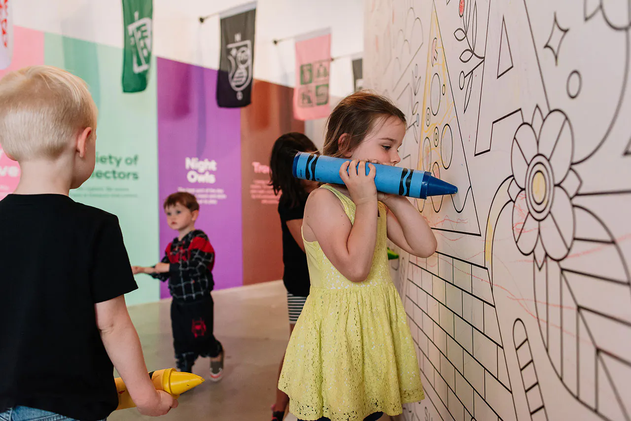 A little girl in a yellow dress holding an oversized blue crayon and coloring on a black and white wall decal.
