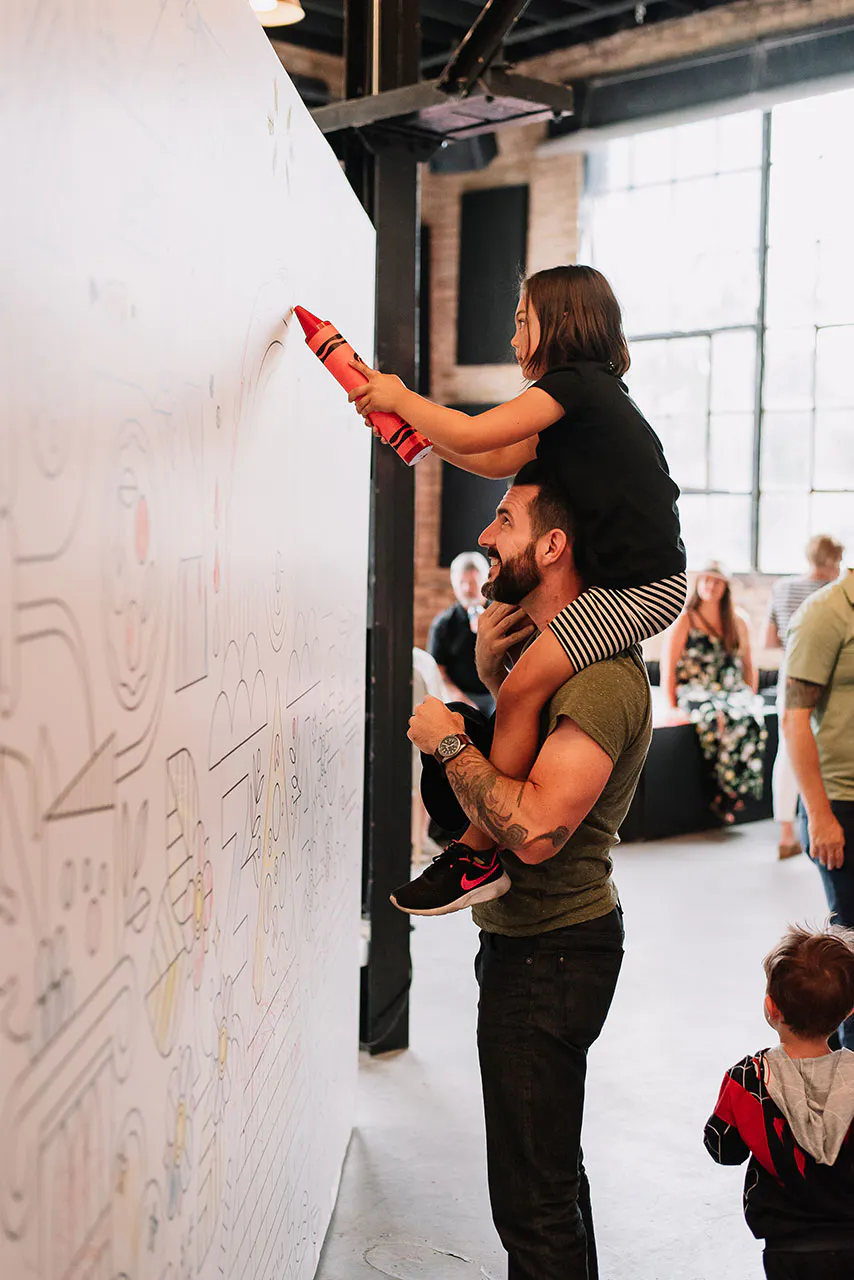A young girl sitting on a man's shoulders and coloring on a wall decal with an oversized orange crayon.