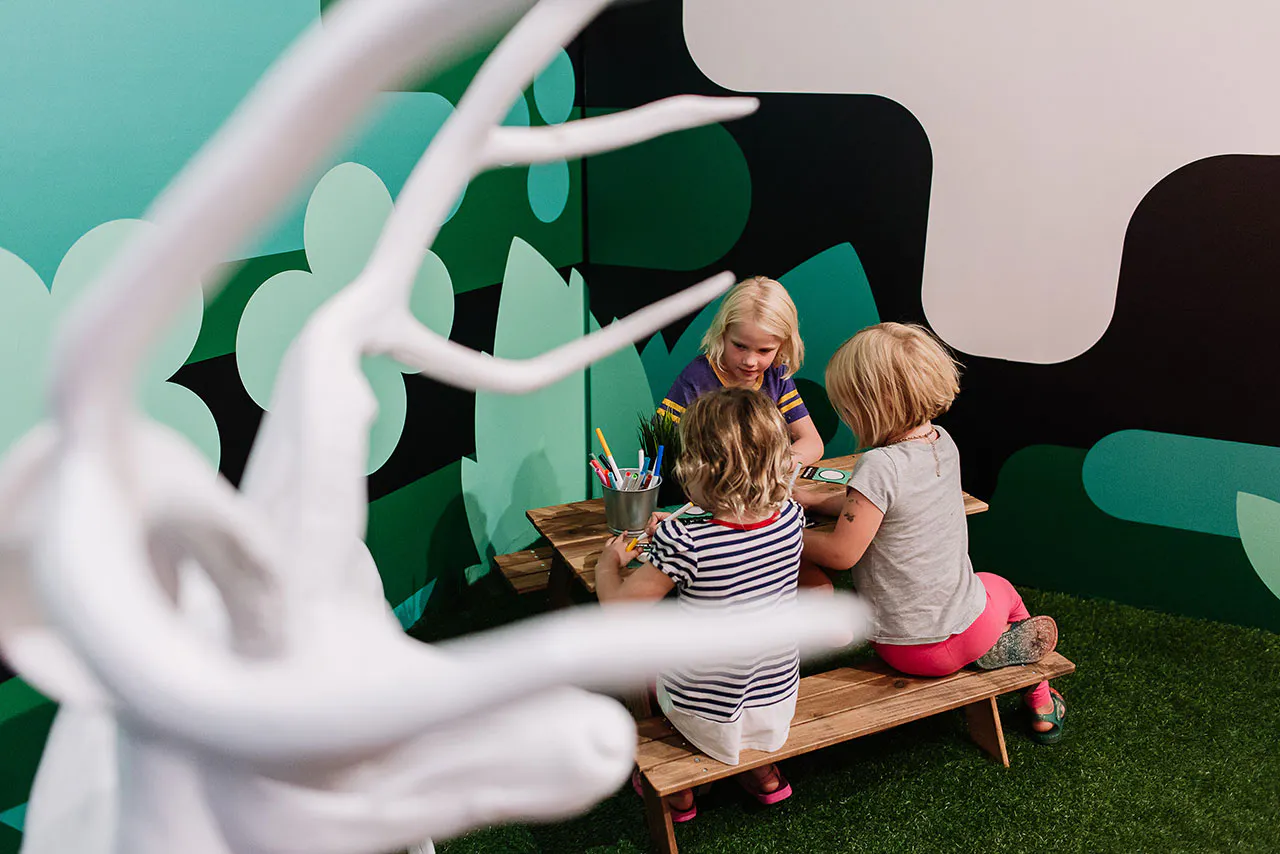 Three children sitting at a small picnic table coloring with a green wall decal behind them.