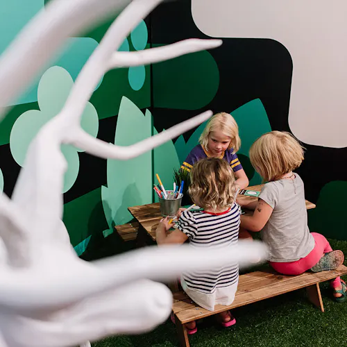 Three children sitting at a small picnic table coloring with a green wall decal behind them.