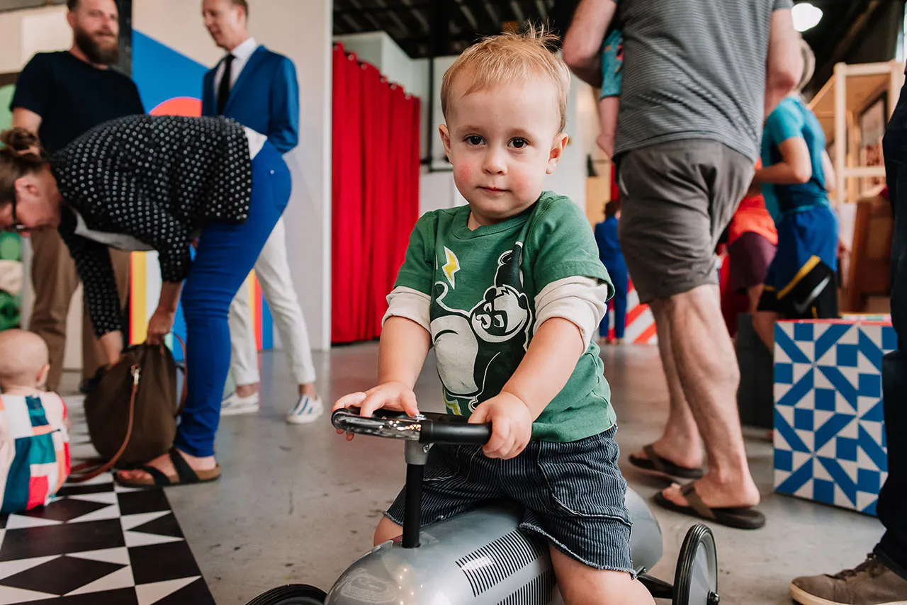 A little boy wearing a green shirt and riding a gray toy tractor with adults around him.