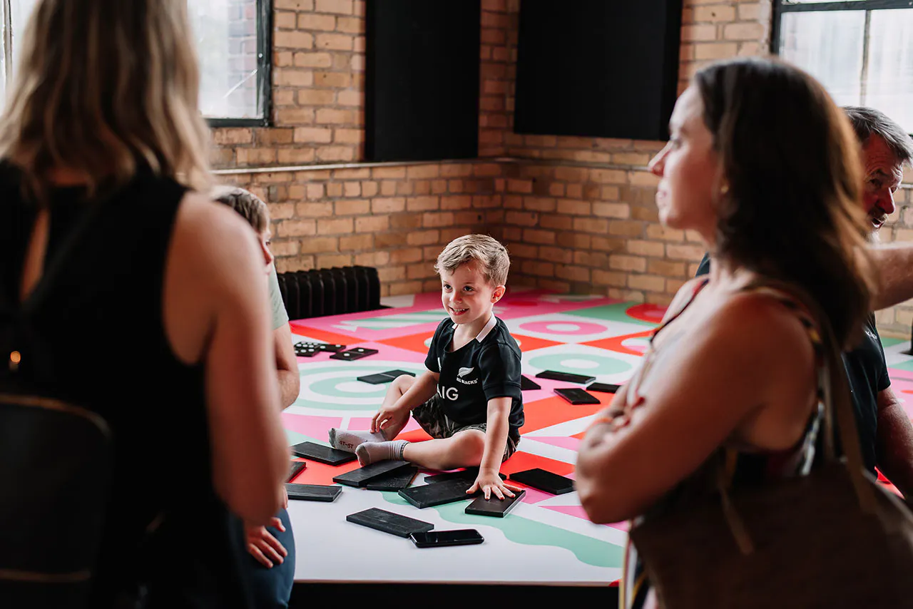 A young boy sitting on a floor covered with a pink, red and teal decal and playing with oversized dominos.
