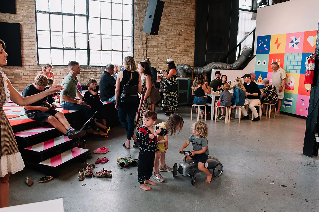 A group of adults and children in Club Kiddo, riding bikes, talking and eating at a table.