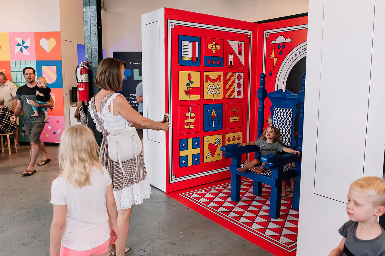 A woman taking a photo of a young boy sitting in a blue chair surrounded by bright red, blue and yellow wall decals.
