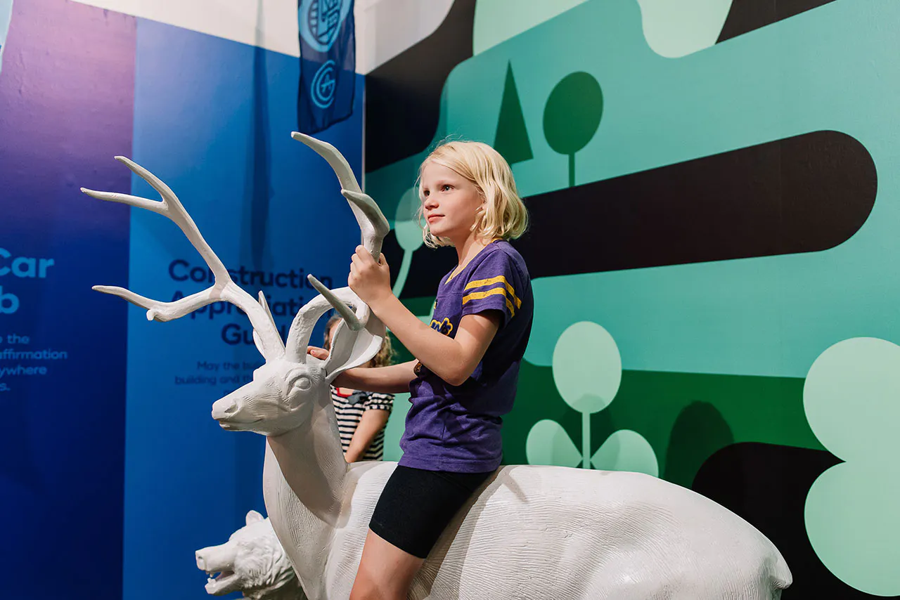 A young girl wearing a purple shirt and black shorts sitting on a white sculpture of a deer.