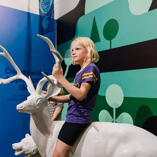 A young girl wearing a purple shirt and black shorts sitting on a white sculpture of a deer.