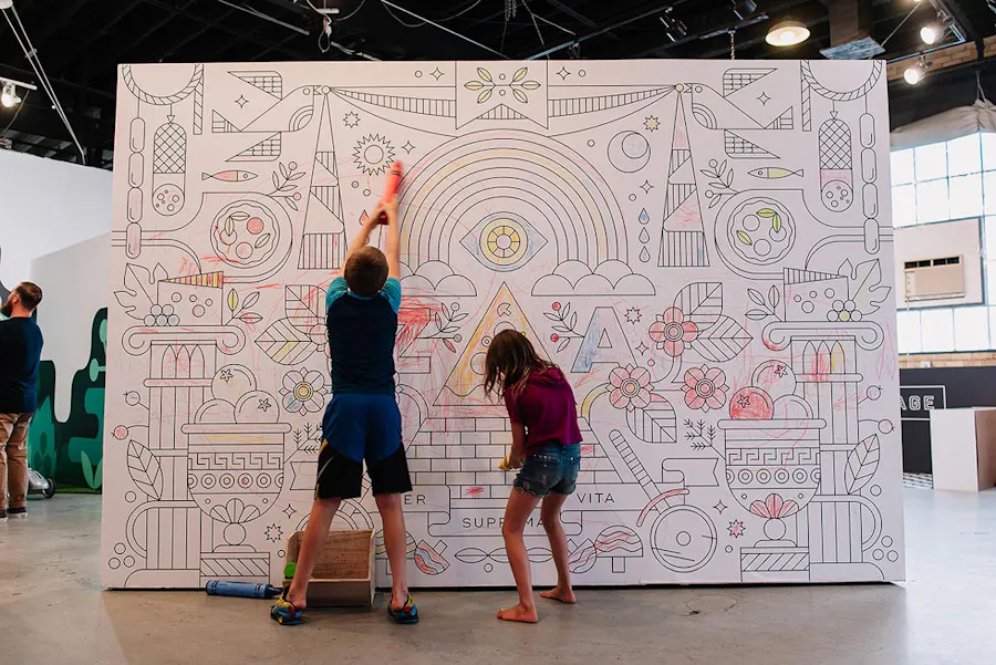 Two kids coloring with big crayons on a custom wall graphic printed with various designs.