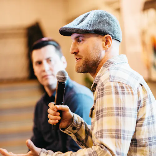Two men sitting on stools giving a presentation, with one man holding a microphone and wearing a hat.