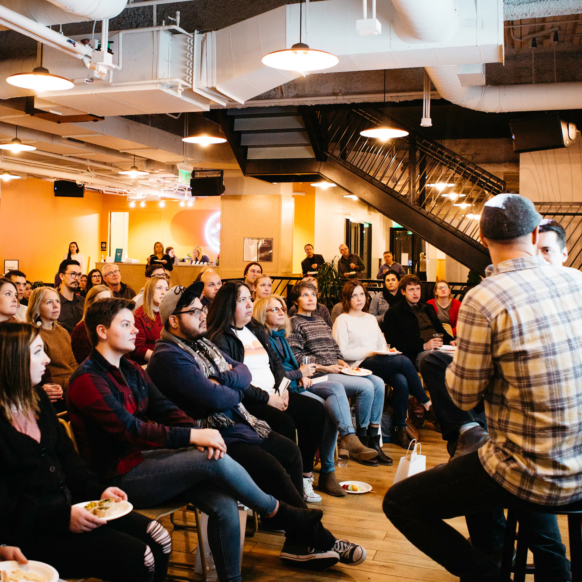Two men sitting on stools talking during a presentation with rows of people sitting in front of them.