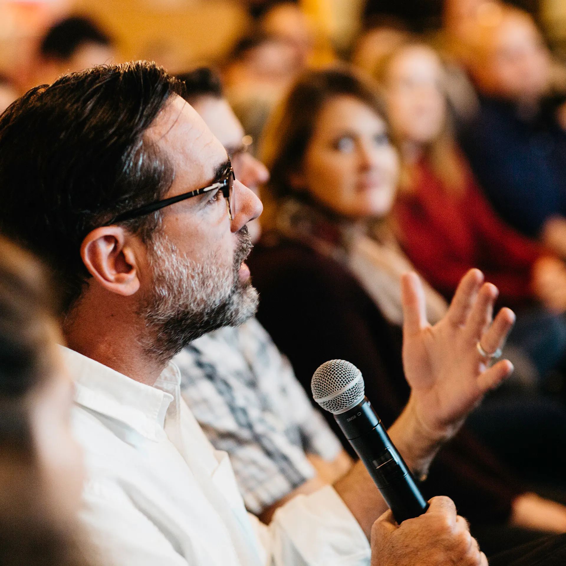 A man in an audience wearing a white shirt and glasses speaking into a microphone at an event.