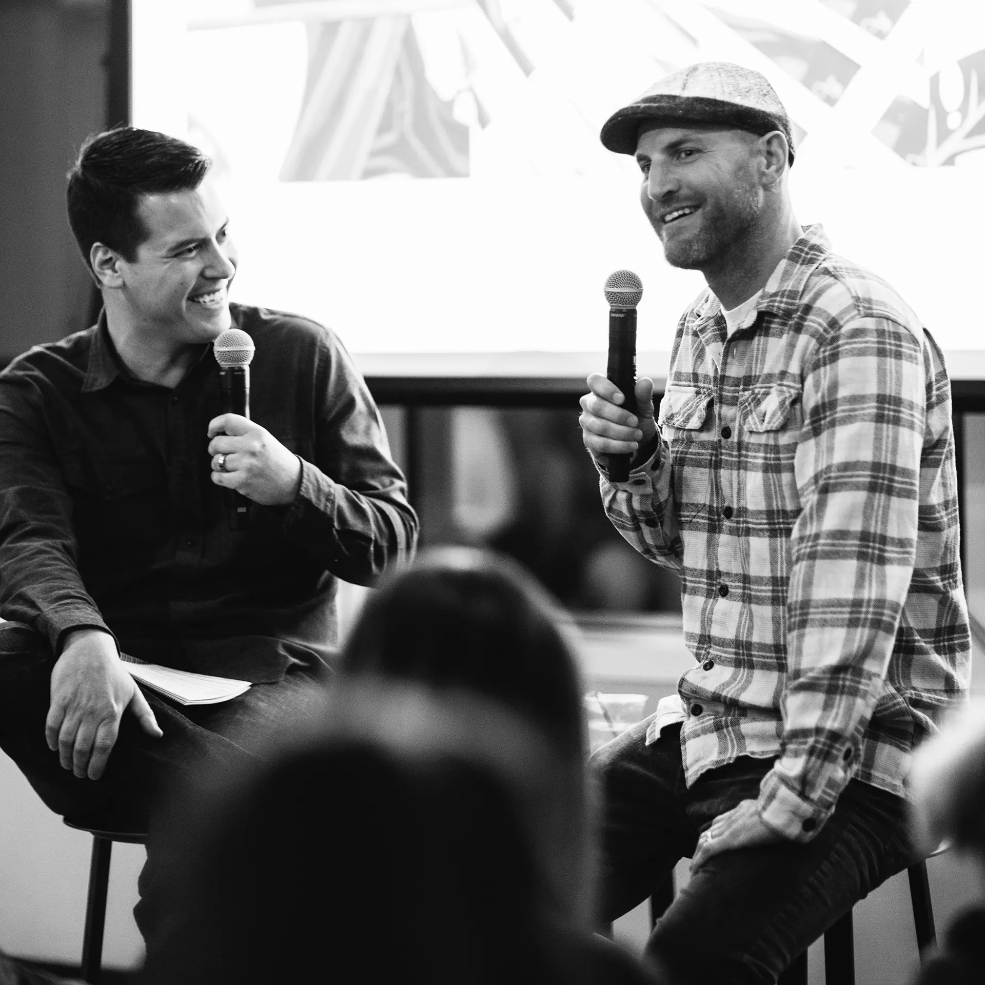 Two men sitting on stools during a presentation, smiling and speaking in front of an audience.