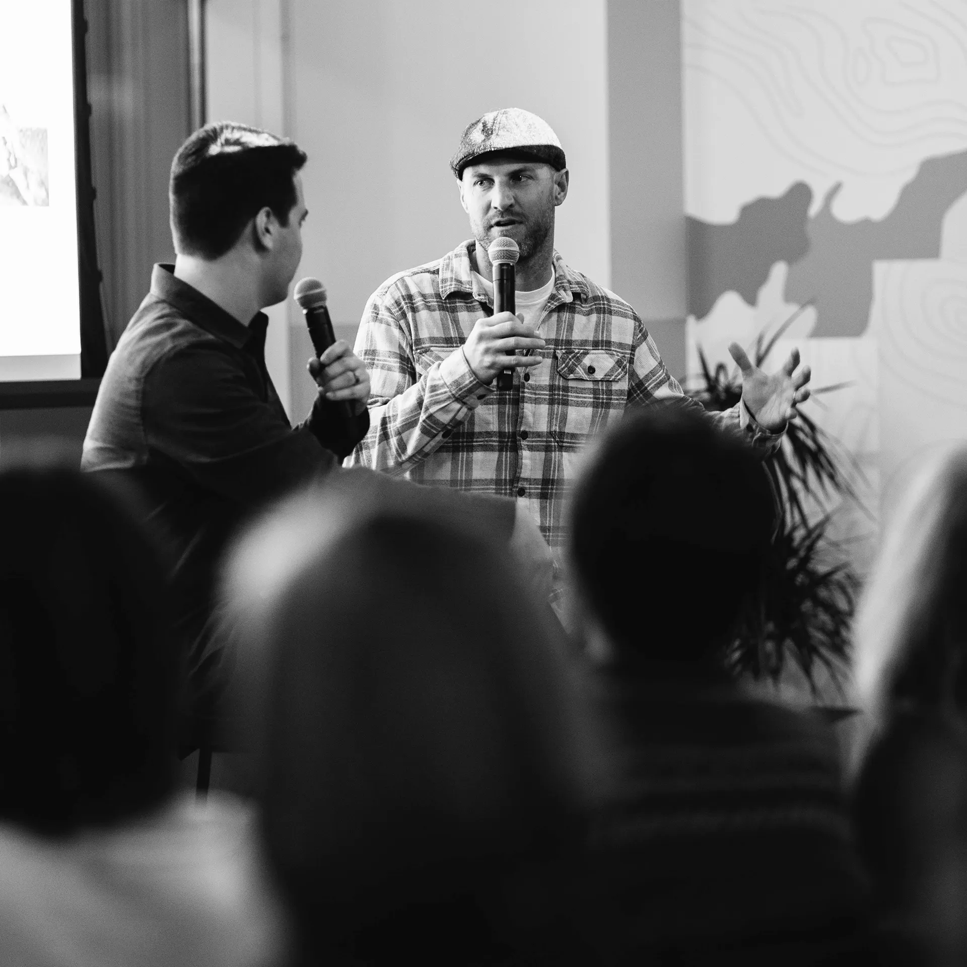 Two men sitting on stools and holding microphones giving a presentation to an audience.